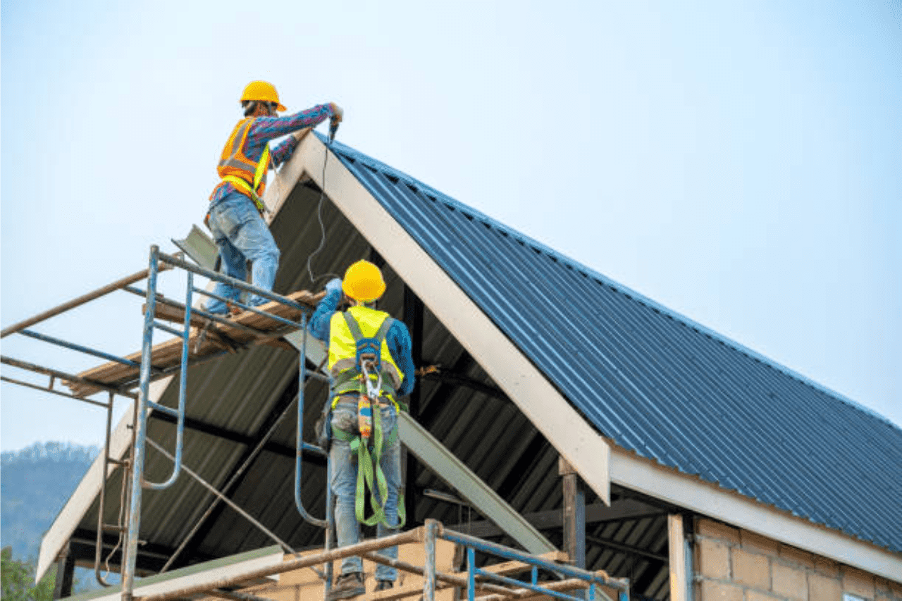 Two roofers in hard hats and safety harnesses fitting metal roof panels from scaffolding